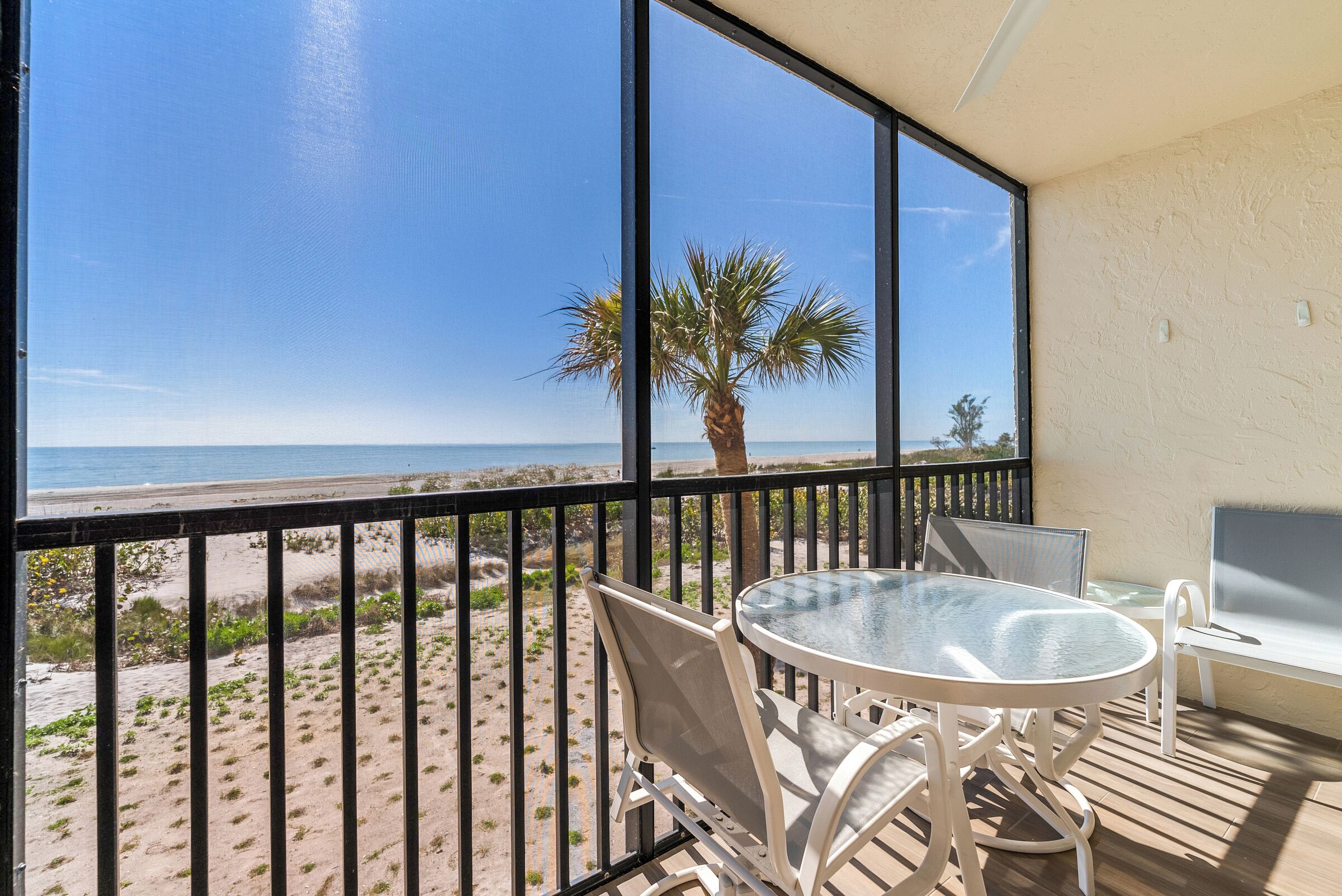 A balcony with a glass table and chairs, overlooking a beach with palm trees and the ocean.