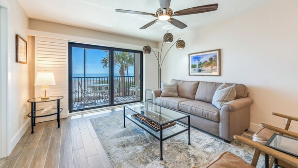 A living room with a glass coffee table, a beige sofa, a ceiling fan, and a view of palm trees through large sliding glass doors. - OCEAN'S REACH 4C2: 2nd Floor, Beautiful Gulf Views w/Easy Beach Access! (Sanibel Island)