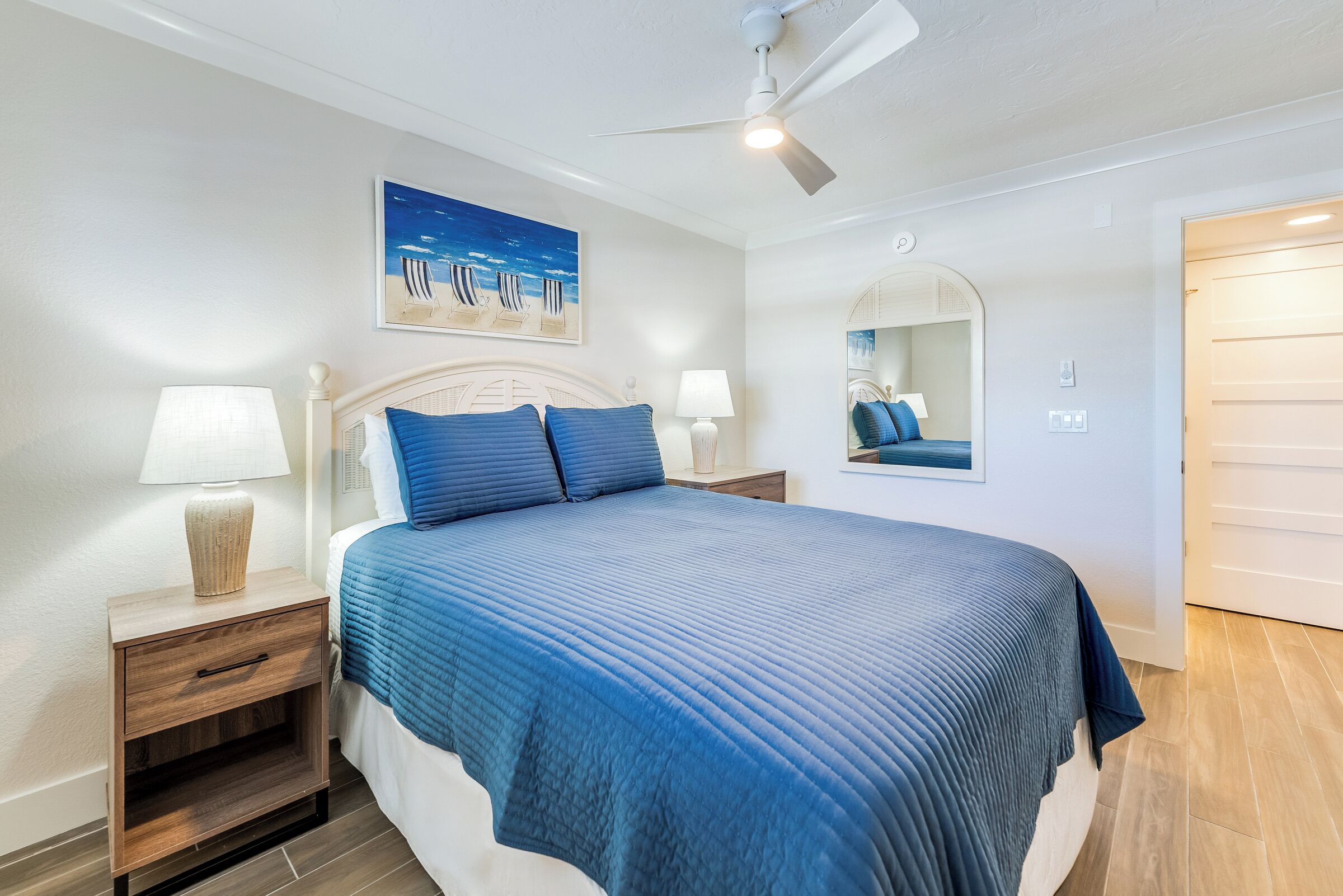A bedroom with a bed, nightstands, a ceiling fan, a painting, and a mirror.