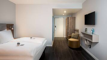 A modern hotel room with a large bed, a TV mounted on the wall, a chair, and a small table with a magazine and a water bottle.