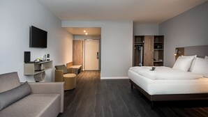 A modern hotel room with a bed, a sofa, a TV, a desk with a coffee maker, and a wardrobe. - elaya hotel oberhausen, Trademark by Wyndham (Oberhausen)