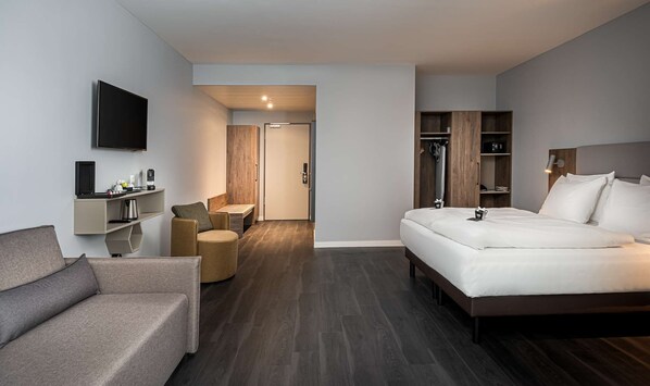 A modern hotel room with a bed, a sofa, a TV, a desk with a coffee maker, and a wardrobe. - elaya hotel oberhausen, Trademark by Wyndham (Oberhausen)