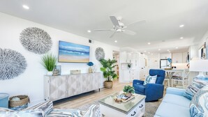 Ein modernes Wohnzimmer mit Sofa, Sessel und Couchtisch. An der Decke hängt ein Ventilator, an der Wand hängt ein Gemälde und steht eine Pflanze.