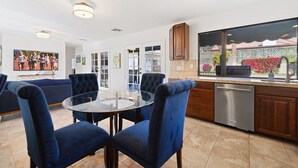 A modern kitchen with a glass dining table, blue upholstered chairs, and a view of a garden through a large window. - Centrally located Palm Desert Oasis (Palm Desert)