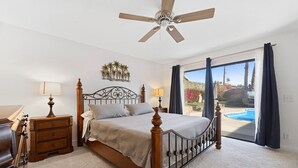 A bedroom with a bed, a ceiling fan, a nightstand, a lamp, and a view of a swimming pool. - Centrally located Palm Desert Oasis (Palm Desert)