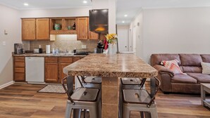 A modern kitchen with wooden cabinets, a granite countertop, and a built-in oven. There is a brown leather couch and two bar stools in the living area.
