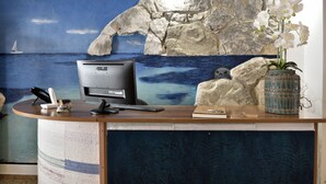 A reception desk with a computer, a vase of white orchids, and a stone wall mural depicting a coastal scene. - Hotel Goloritzé (Baunei)