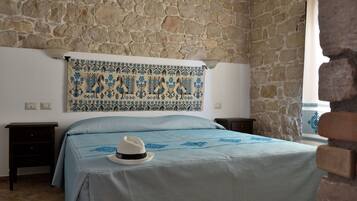 Una habitación con pared de piedra, una cama con colcha azul, un tapiz decorativo y un sombrero blanco sobre la cama.