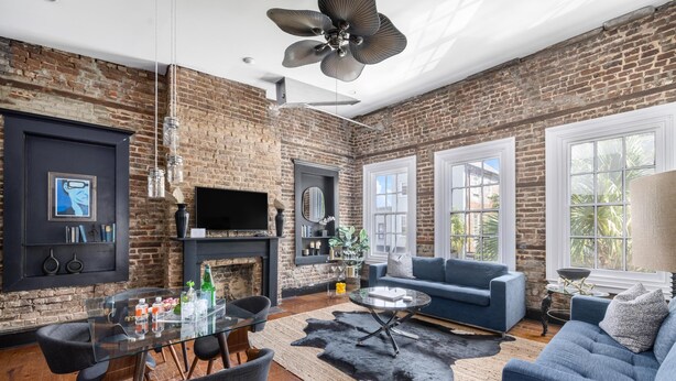 Una sala de estar moderna con pared de ladrillo, chimenea, sofá azul, mesa de comedor de vidrio y ventilador de techo.