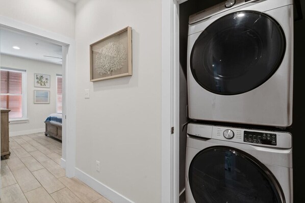A laundry room with stacked front-loading washers and dryers, a framed artwork on the wall, and a glimpse of a bedroom with a bed and window blinds. - Hammock Dunes I - Private Cottage Beach Side (Orange Beach)