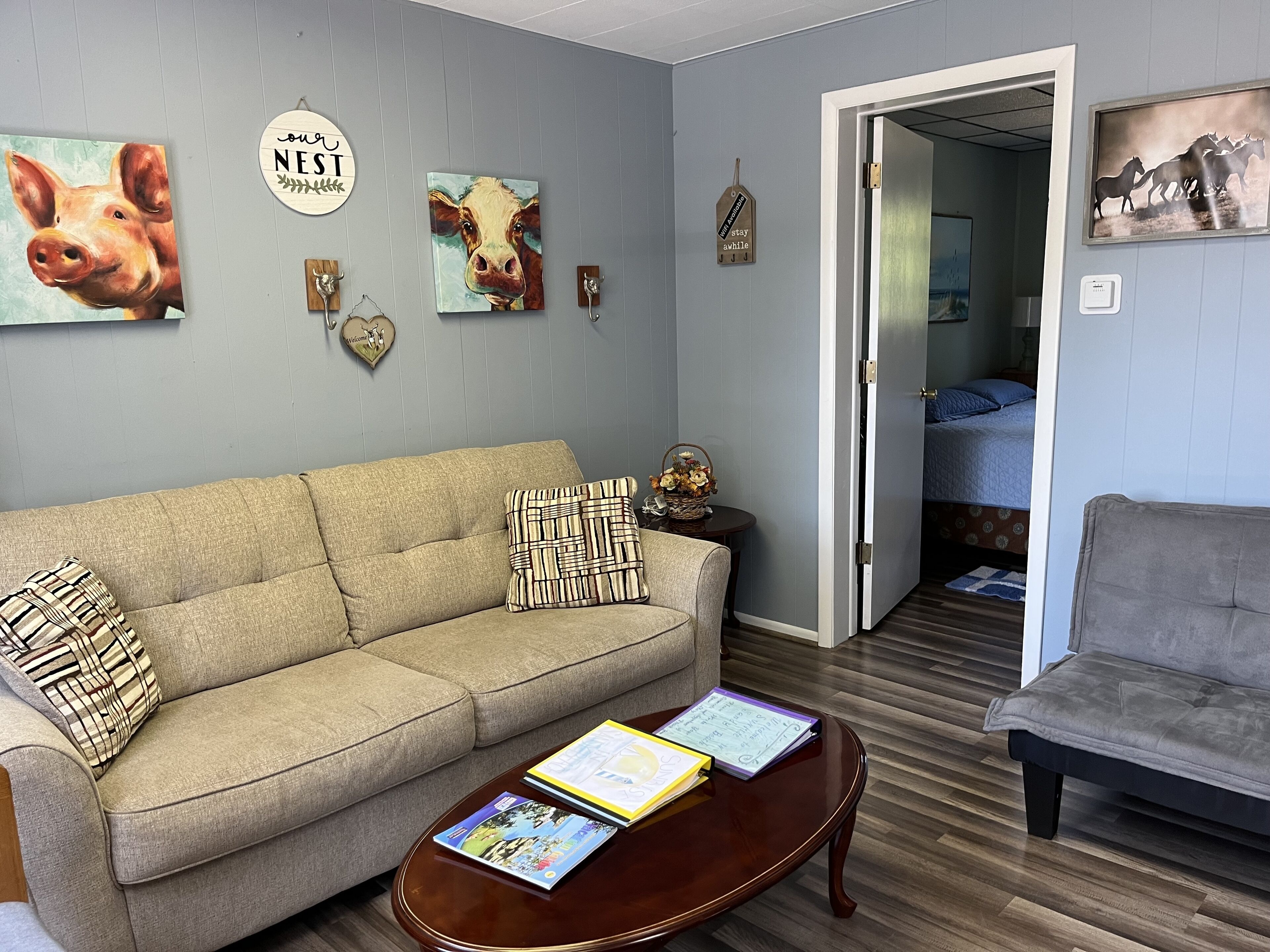 Ein Wohnzimmer mit einem beigen Sofa, einem grauen Sessel und einem Couchtisch mit Zeitschriften. An den Wänden befinden sich Dekorationen, darunter ein Gemälde von einer Kuh und ein Schild mit der Aufschrift 'Our Nest'.