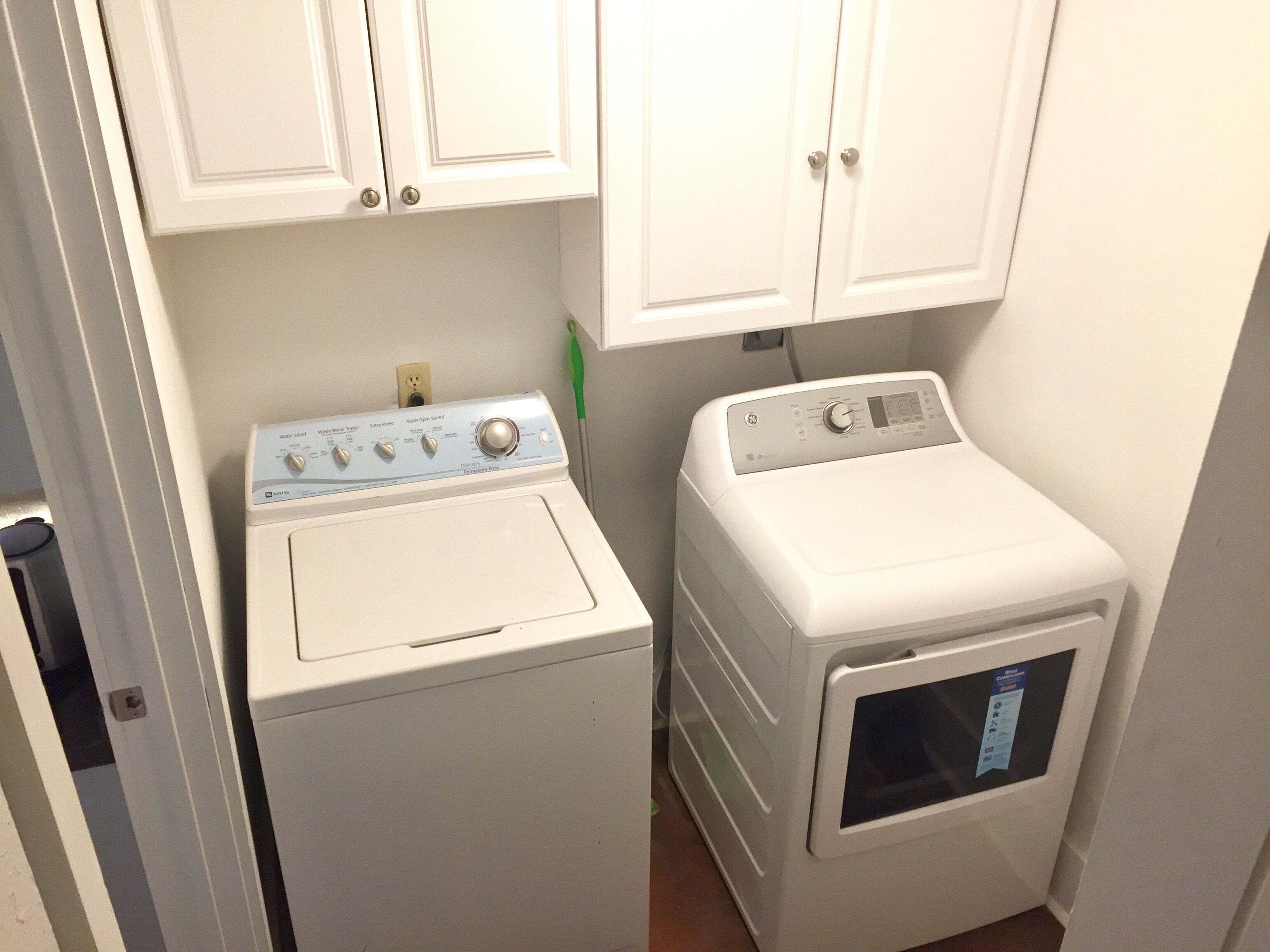 A laundry room with a front-loading washer and dryer, white cabinets above, and a green bottle on the floor.