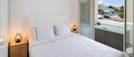 A bedroom with a large bed, a nightstand with a lamp, and a view of a pool and cars outside.