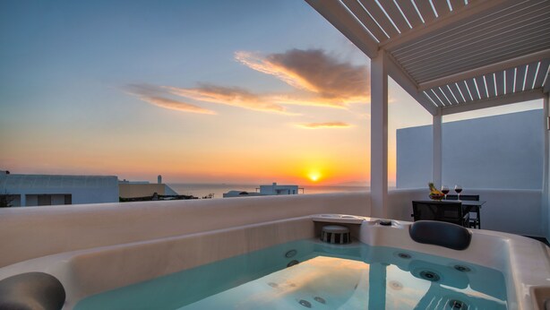 Un jacuzzi en la azotea con vistas al atardecer, una mesa con dos copas de vino y una planta pequeña.
