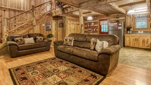 Een rustiek houten interieur met een wenteltrap, twee leren banken met decoratieve kussens, een kamerbreed tapijt en een keuken met houten kasten en een koelkast.