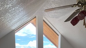 A room with a high ceiling, a ceiling fan, a large window with a view of a landscape, and a framed tapestry depicting a deer. - Torrey Pines Cozy Cabin! (Torrey)