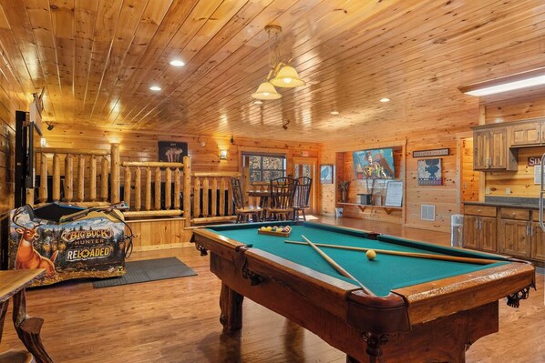 A wooden interior with a pool table, a deer-themed arcade game, and a rustic wooden wall.