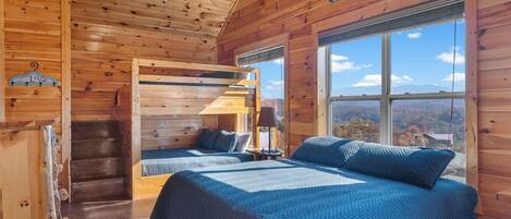 A wooden cabin bedroom with two beds, a window with a mountain view, and a ladder leading to a loft area.