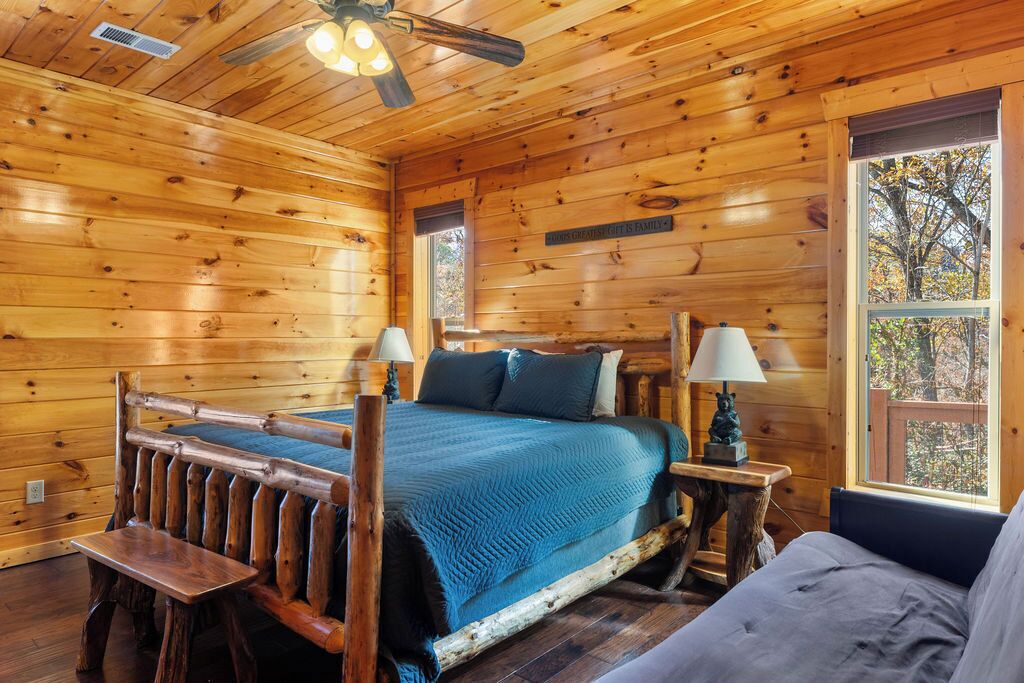 A wooden cabin bedroom with a bed, two bedside tables with lamps, a bench, and a window with a view of trees.