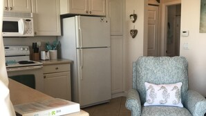 A compact kitchen with white cabinets, a microwave, a refrigerator, and a stove. There's a patterned armchair with a decorative pillow. - Amazing one bedroom direct ocean front condo with beautiful view of the ocean! (COCOA BEACH)