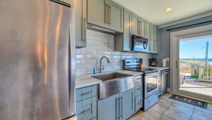 A modern kitchen with stainless steel appliances, grey cabinets, and a view of the beach through a sliding glass door. - SeaClusion South - Oceanfront - Dog Friendly (Topsail Beach)
