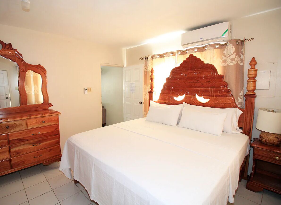 Un dormitorio con cabecera de madera, cómoda de madera, mesita de noche con lámpara y equipo de aire acondicionado sobre la cama.