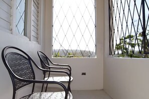 A balcony with two black metal chairs, a window with a diamond-patterned metal screen, and a view of greenery and hills.