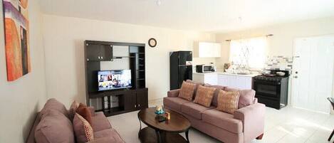 A living room with a sofa, a coffee table, and a TV. There is a kitchen area with a stove and cabinets.