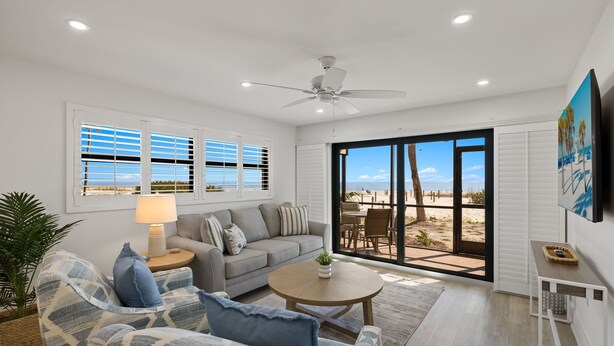 A spacious living room with a sofa, armchair, and coffee table, a ceiling fan, and a view of the beach through large sliding doors.
