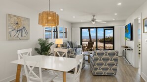 A spacious living area with a dining table, white chairs, a sofa, and a large window offering a view of the beach.