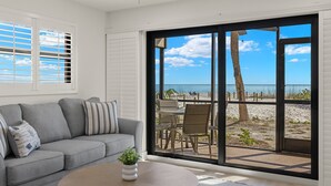 A room with a grey sofa, a round table, and a view of a beach through large glass doors.