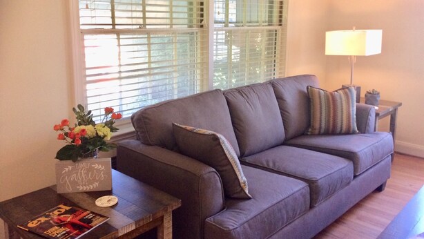 Ein Wohnzimmer mit grauem Sofa, einem kleinen Holztisch mit einer Zeitschrift, einer Blumenvase und einer Lampe.