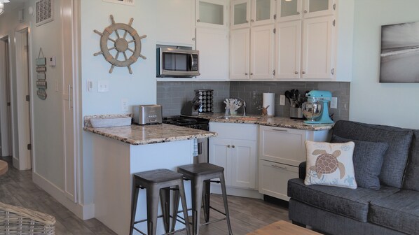 A kitchen with white cabinets, a microwave, a toaster, and a coffee maker. There is a bar stool and a gray couch with a turtle pillow. - Amazing Front Facing Top Floor with unobstructed Gulf and Beach View!! #302 (Galveston)