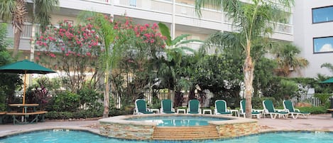 A swimming pool with lounge chairs and a stone border, surrounded by lush greenery and a multi-story building with balconies.