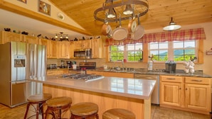 Une cuisine spacieuse avec des meubles en bois, un grand îlot central doté de tabourets, des appareils électroménagers en acier inoxydable et une vue sur la verdure environnante.