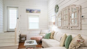A cozy room with a white wooden floor, a white sofa with green and pink cushions, a small table with a pink fluffy stool, and a window with a view of the countryside. - Pigs Fly in Burton just minutes to Round Top Antiques Shopping and Dining (Burton)