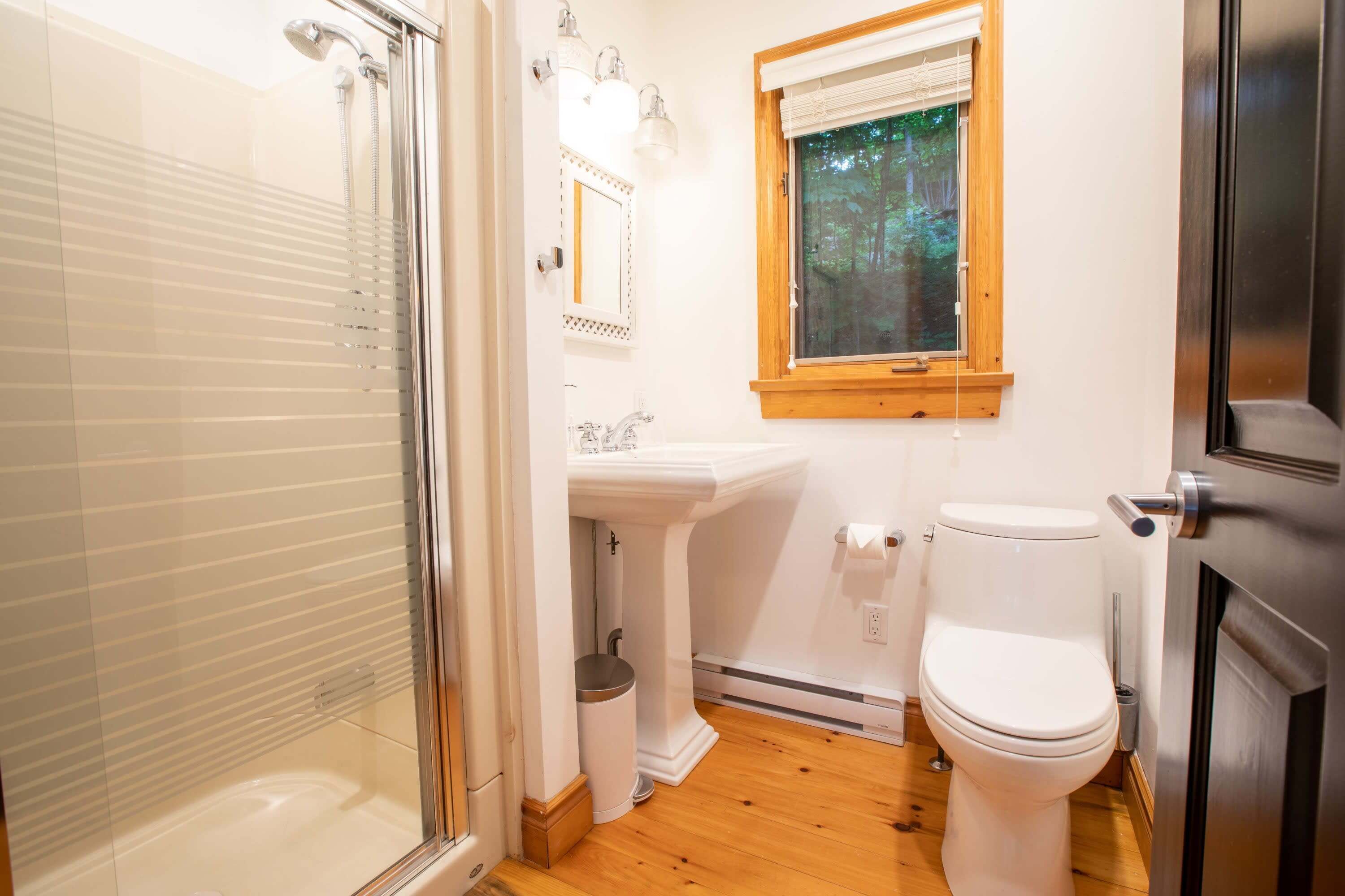 A bathroom with a glass shower enclosure, a sink with a mirror, a toilet, and a window with a view of trees.