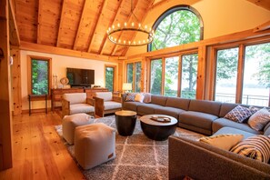 A spacious living room with a large sectional sofa, ottomans, and a coffee table. A television is mounted on the wall, and there are large windows providing natural light and views of the outdoors.