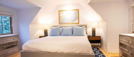 A bedroom with a large bed, two bedside tables with lamps, a dresser, and a window with blinds.