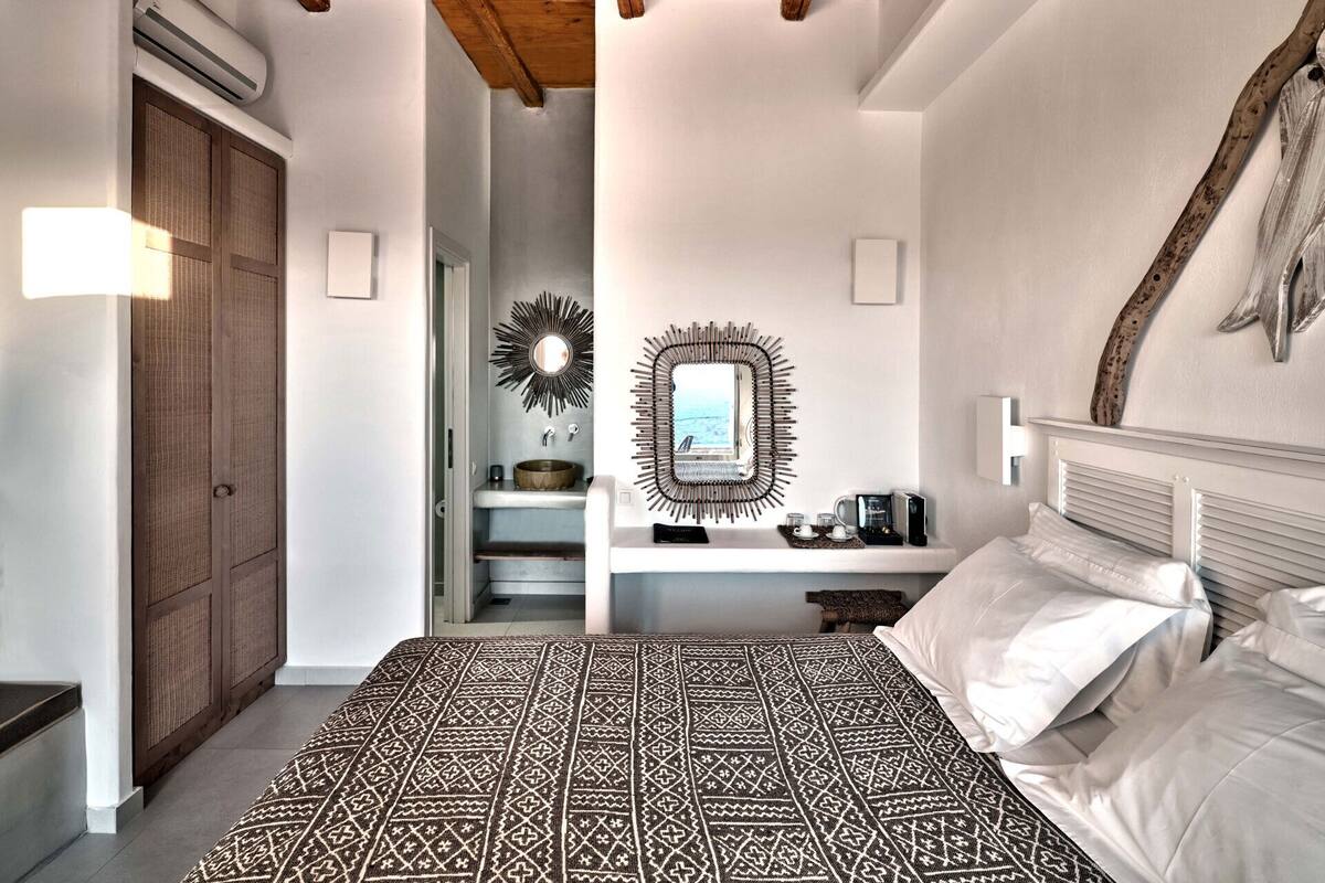 A bedroom with a patterned bedspread, a wooden ceiling, a white bedside table with a mirror, and a wooden door.