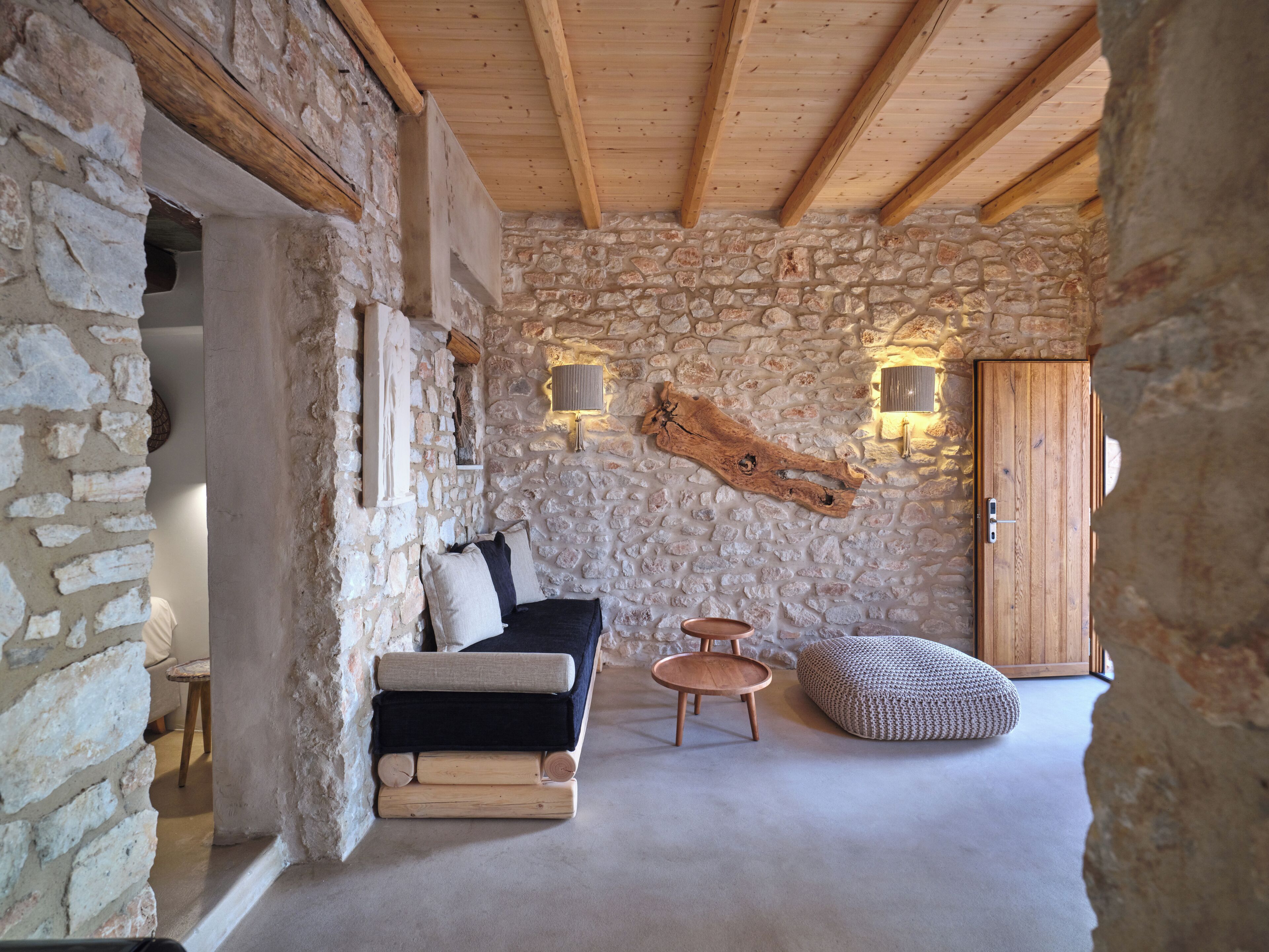 A rustic stone interior with a wooden ceiling, a sofa, a round wooden table, and a wooden cabinet.