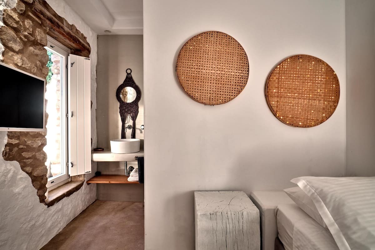 A bedroom with a stone wall, a white bed, a bedside table, a round woven wall hanging, and a white bedside lamp.