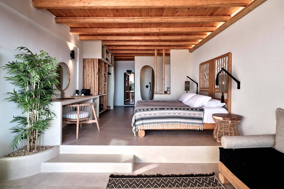 A bedroom with a wooden ceiling, a bed with patterned bedding, a bedside table, a chair, and a large potted plant.