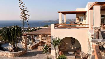 A Mediterranean-style villa with a terrace overlooking the sea, featuring stone walls, a wooden pergola, and outdoor seating.
