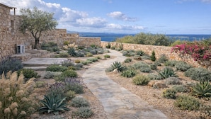 Un chemin de pierres qui traverse un jardin où poussent différentes espèces de plantes, le long d’un mur de pierres.