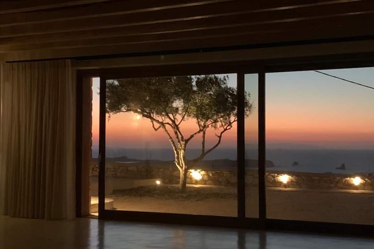 A room with a large window overlooking a sunset, a tree, and a beach with lights.