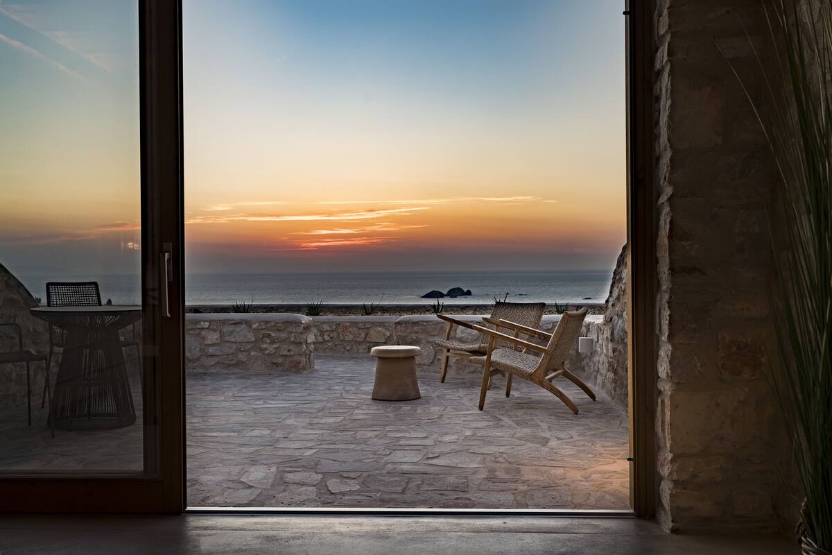 An open patio with stone flooring, wicker furniture, and a view of the sea at sunset.