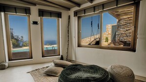 Une pièce dotée de grandes fenêtres donnant sur une piscine et un mur en pierre, et comprenant un pouf en fibres tressées et une petite table.
