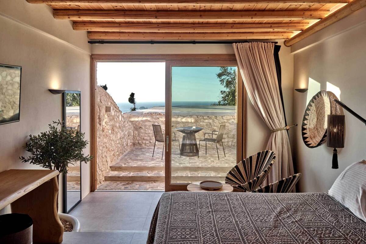 A bedroom with a large bed, a wooden desk, a potted plant, and a view of the sea through a glass door.