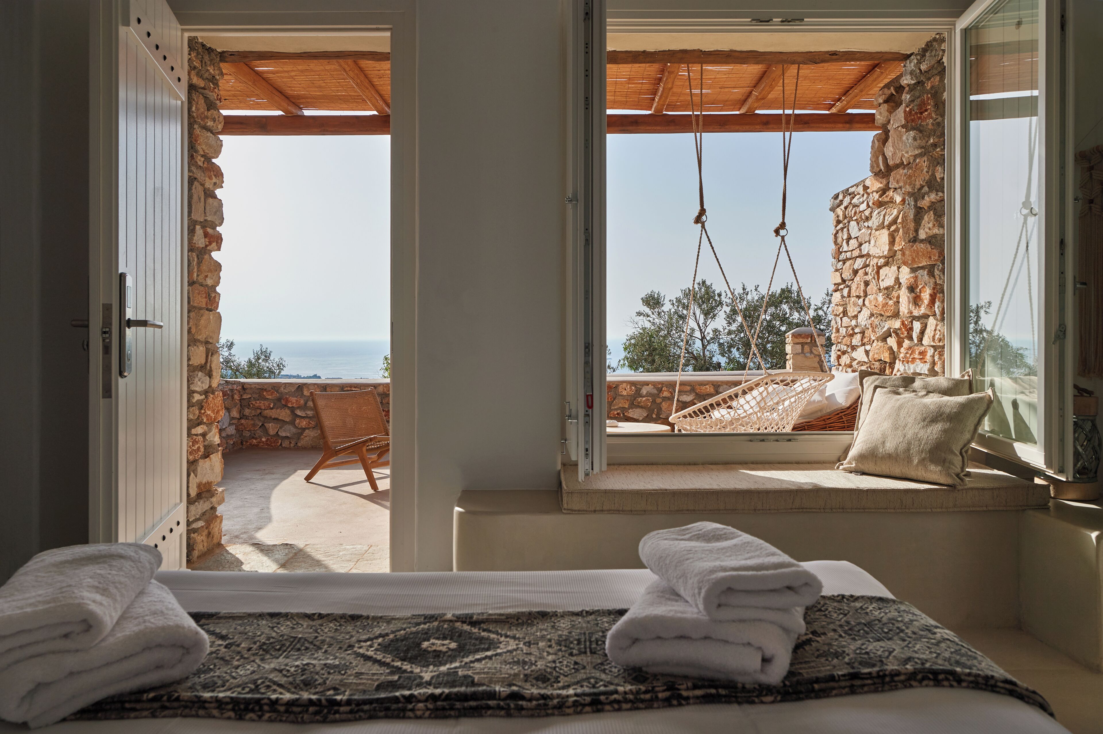 A room with a bed, a view of the sea, a stone wall, and a wooden ceiling.